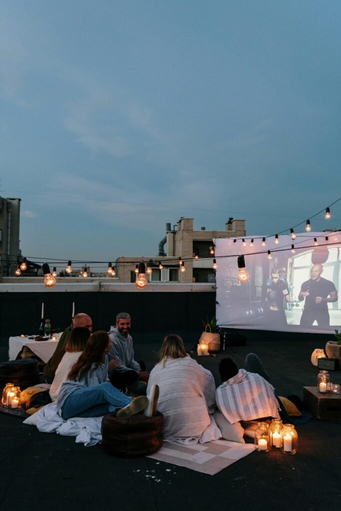 Home 5 A group of friends enjoying an outdoor movie on a rooftop at dusk, surrounded by lights.