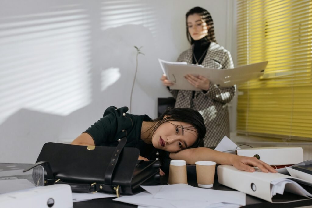 What to Do About Night Shift Memory Problems and Cognitive Decline 7 Exhausted employee resting on desk surrounded by documents, colleague in background.