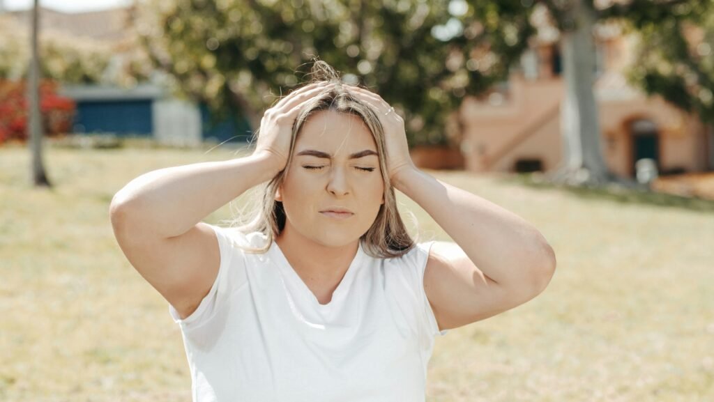 What to Do About Night Shift Memory Problems and Cognitive Decline 2 Adult woman holding her head in pain, sitting outdoors under sunlight, likely experiencing a migraine.