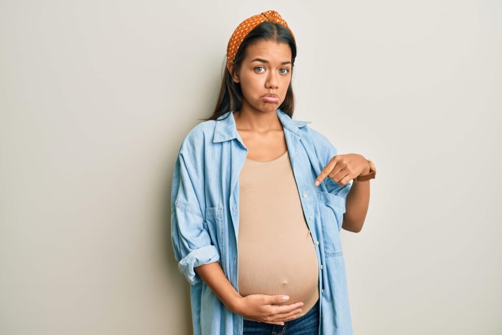 beautiful hispanic woman expecting baby touching pregnant belly pointing down looking sad upset indicating direction with fingers unhappy depressed 1