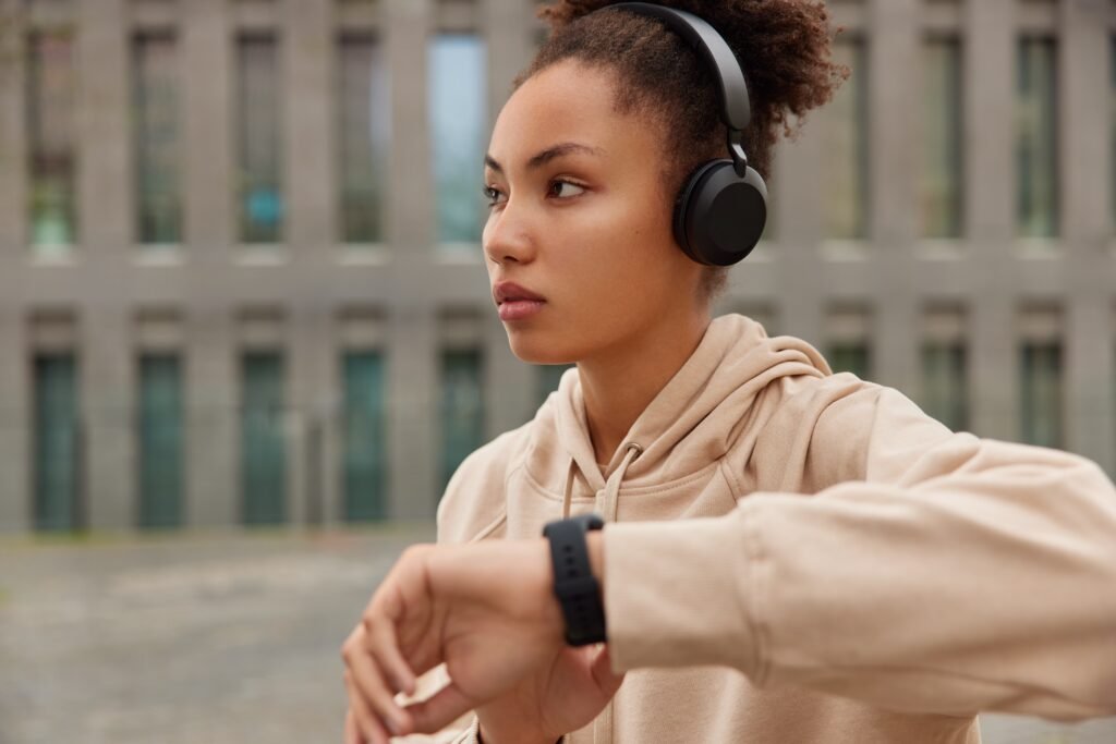 Night Shift and Wearables: How to Use a Fitness Tracker to Actually Improve Your Health 2 thoughtful fitness woman checks results cardio training smartwatch focused away wears casual hoodie listens music via headphones poses outdoors near building sporty lifestyle concept 1